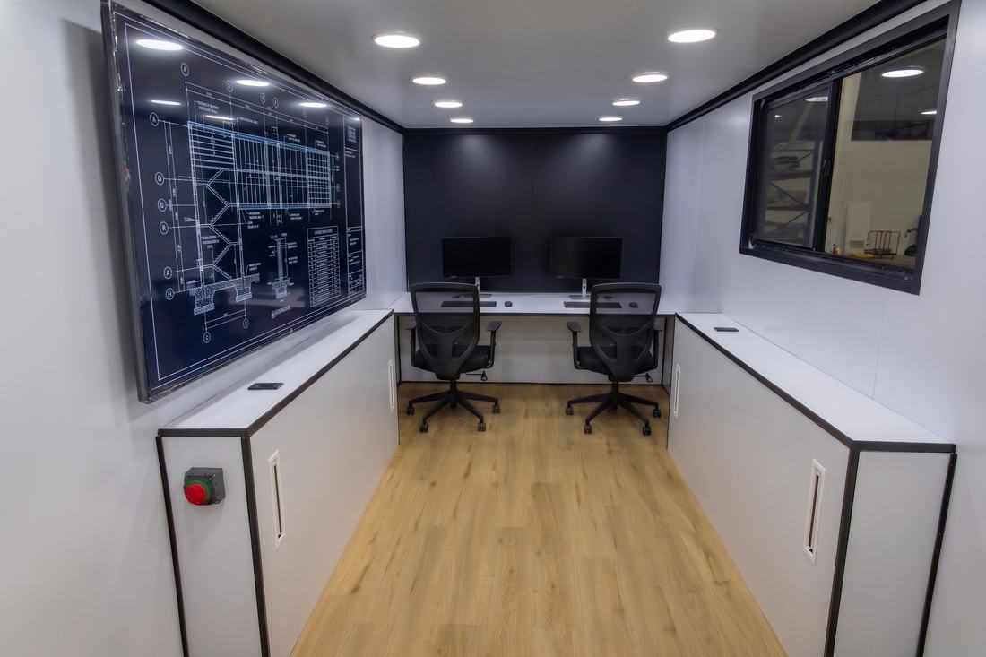 Interior of a SPOG trailer set up as a jobsite office with two workstations, monitors, a wall-mounted plan display, and recessed lighting.