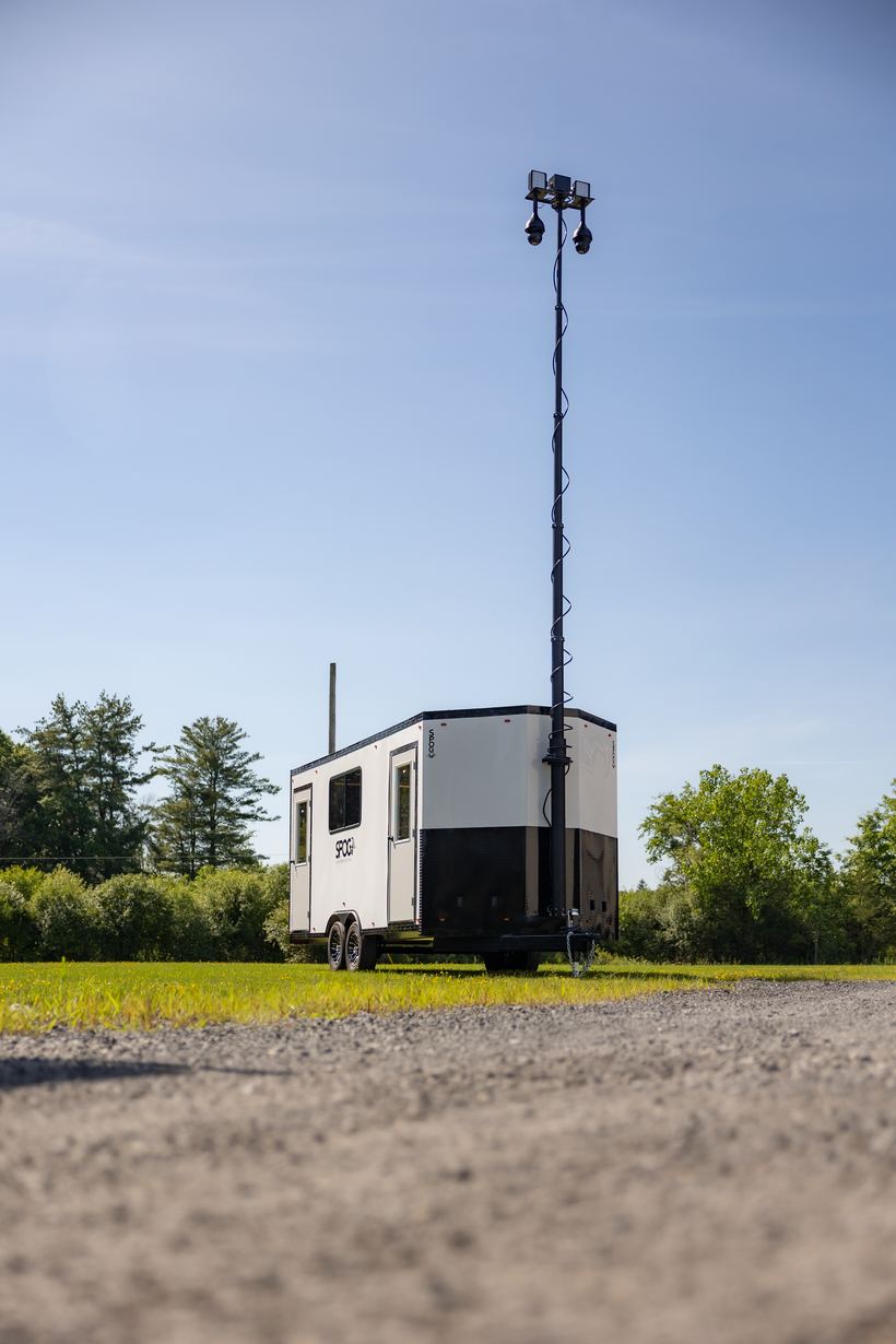 A SPOG trailer parked on a grass lot with its telescoping tower fully extended, carrying floodlights and two pan-tilt-zoom jobsite cameras high above the roofline.