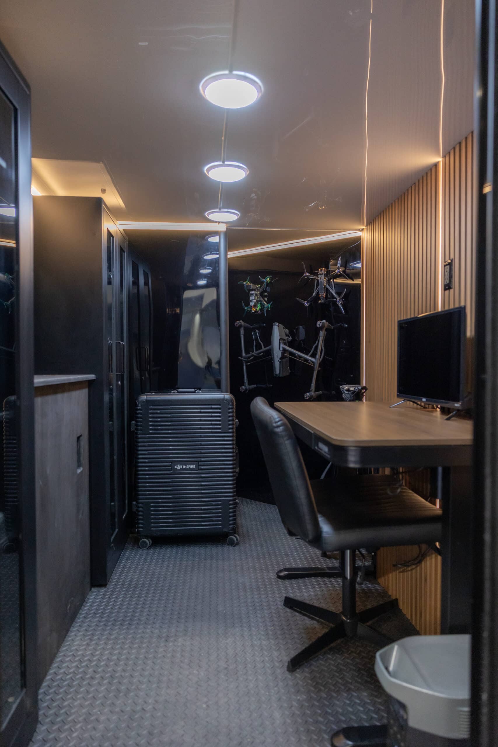 Interior of a modern solar-powered trailer with a wooden finish, featuring a wall-mounted screen playing a scene from The Matrix, a cozy built-in bench, minimalist desk setup, and overhead lighting creating a warm ambiance.
